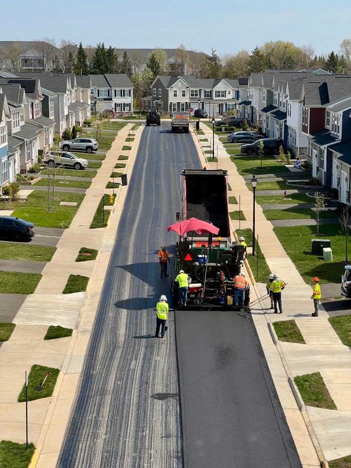 Workers repaving a street in a suburban residential neighborhood on a sunny day.